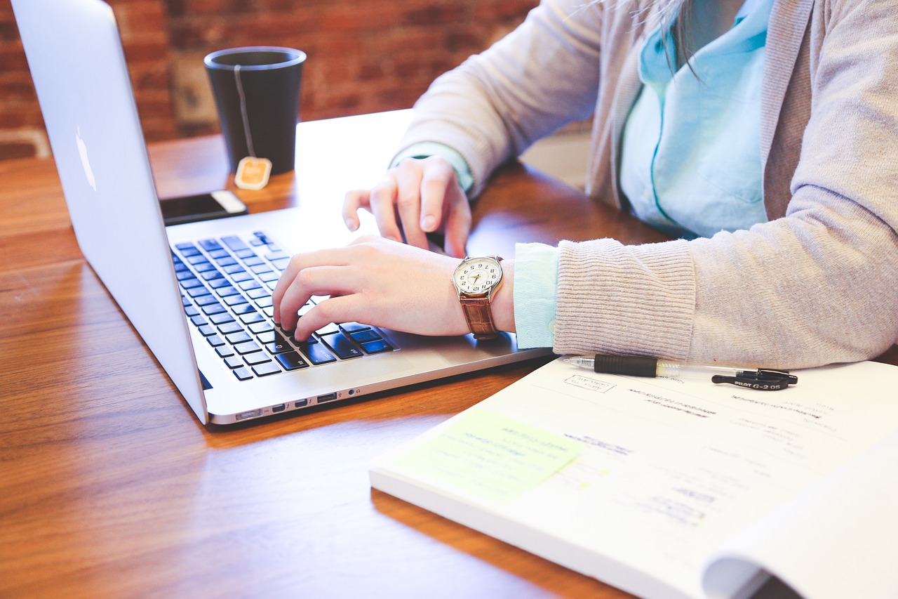 Woman drinking tea doing office work on a laptop with cellphone and book.