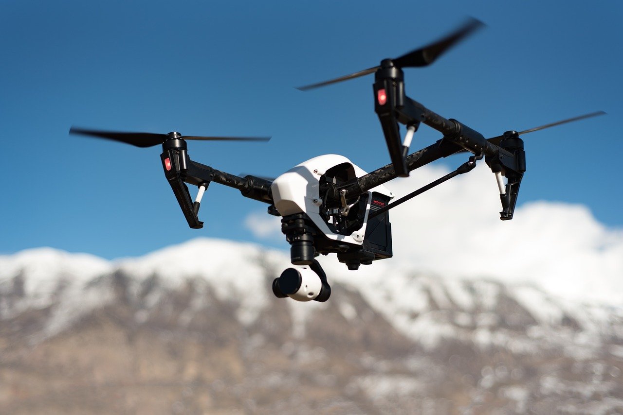 A white drone flying in a blue sky, snowcapped mountains below, white puffy white clouds above them, the drones rotorts akimbo whirling, camera extended below watching something off camera.