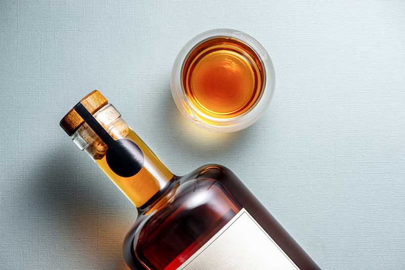 A light colored background, top-down, with a blank-label whiskey bottle and an amber liquid poured into a glass.