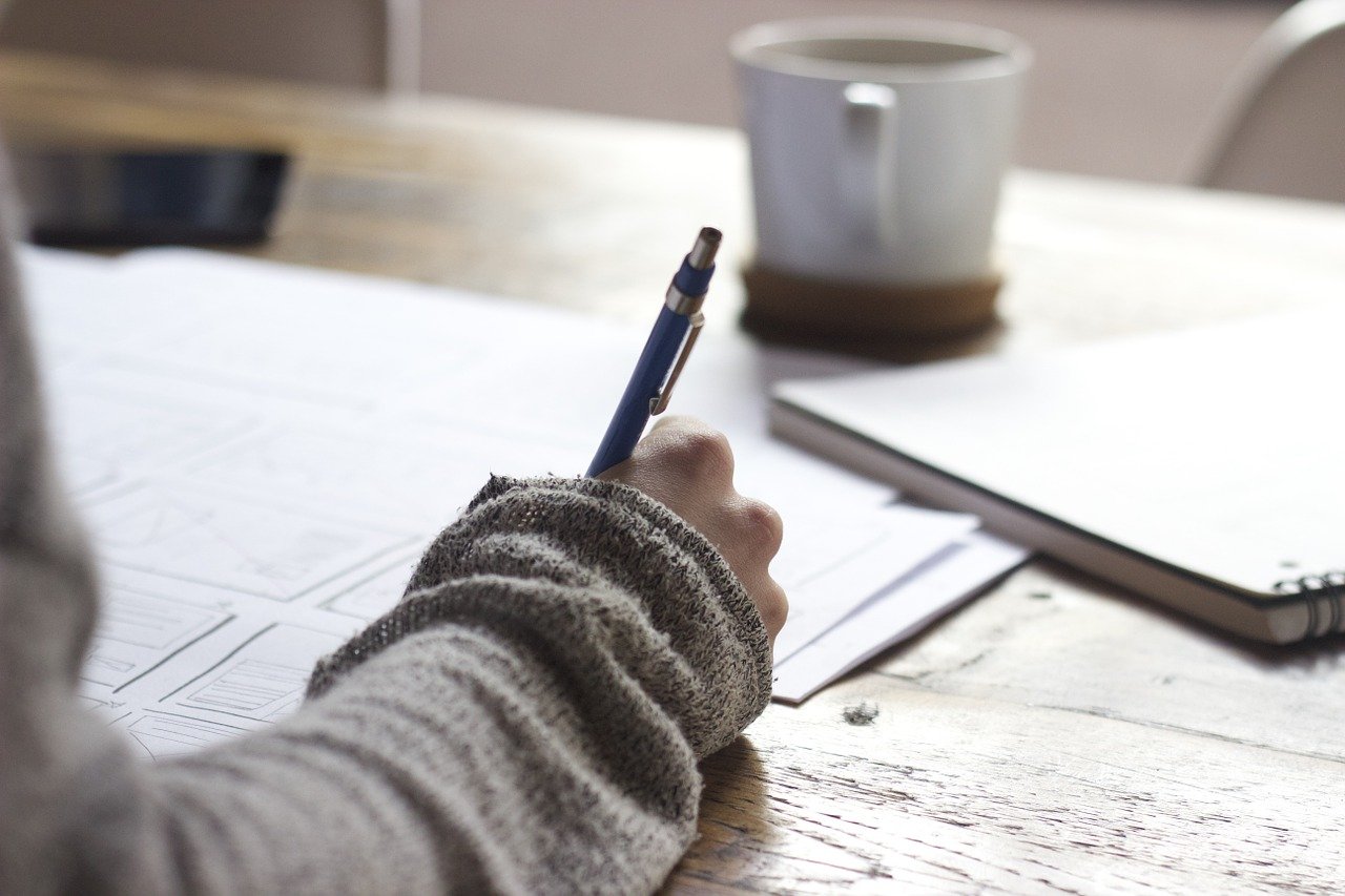 Papers laid out on a wooden table, person's arm visible, dark writing pen visible, writing, wearing a tan, wrinkled, itchy-looking sweater, white, thick cup of hot cocoa sits on a coaster further away slightly out of reach.