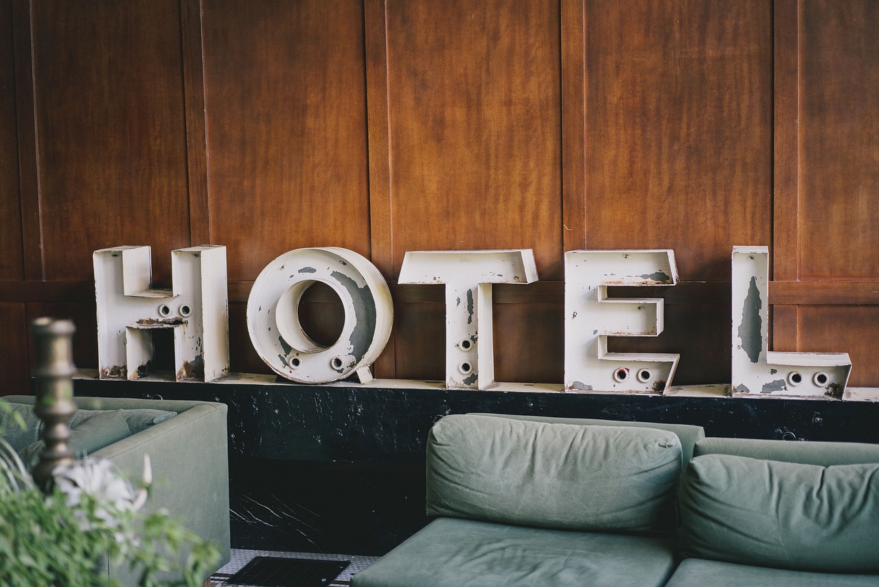 A hotel lobby with a beige couch, grey, poofy pillows, and an industrial metal cutout "H O T E L" against the wall.