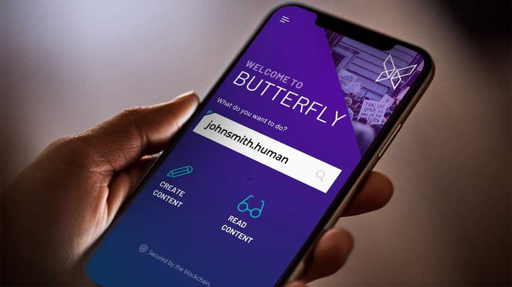 Hand holding a smartphone with the Butterfly Protocol Dapp visible on the screen, a purple user interface displays words "Welcome to BUTTERFLY: What do you want to do johnsmith. human?"