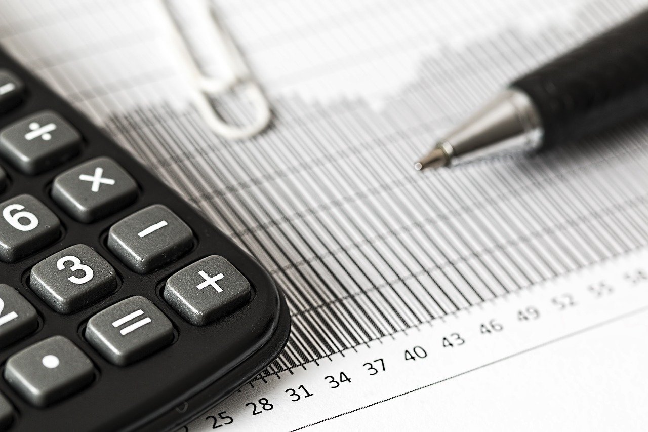 A calculator, white paperchip, and ballpoint pen lay atop a barchart on a white table