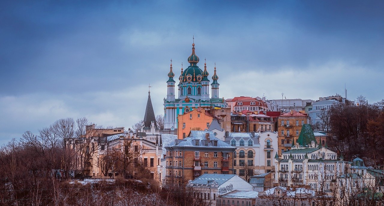 A steepled building in Kyiv city Ukraine it pokes out amid other different colored buildings, some blue, orange, white, the architecture is old and resplendent.