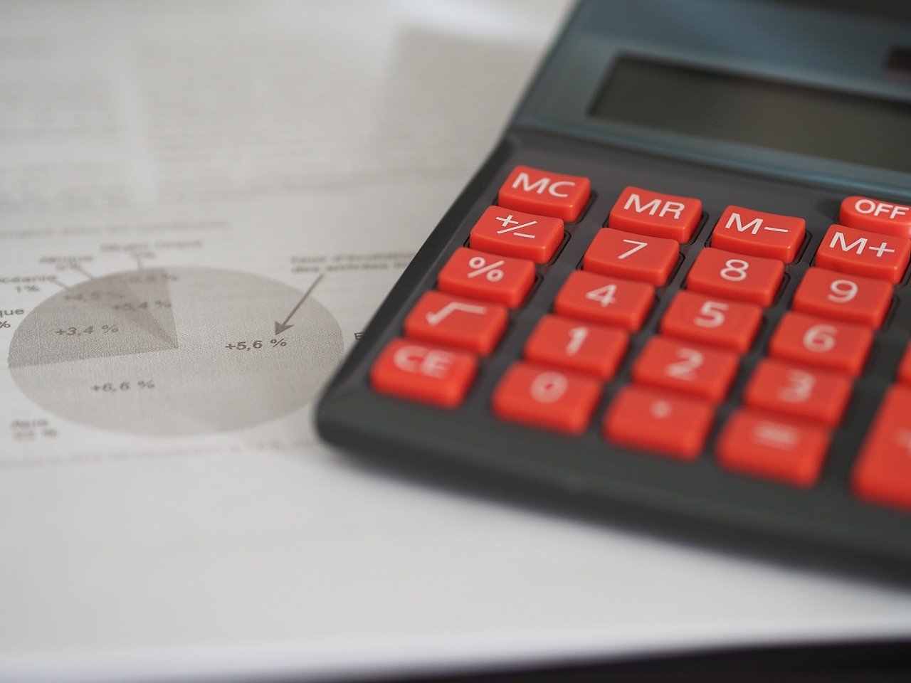 A calculator with huge red buttons in a black and white photograph sitting on top of greyscale documents showing mathematical figures such as pie charts, numbers and other financial facts.