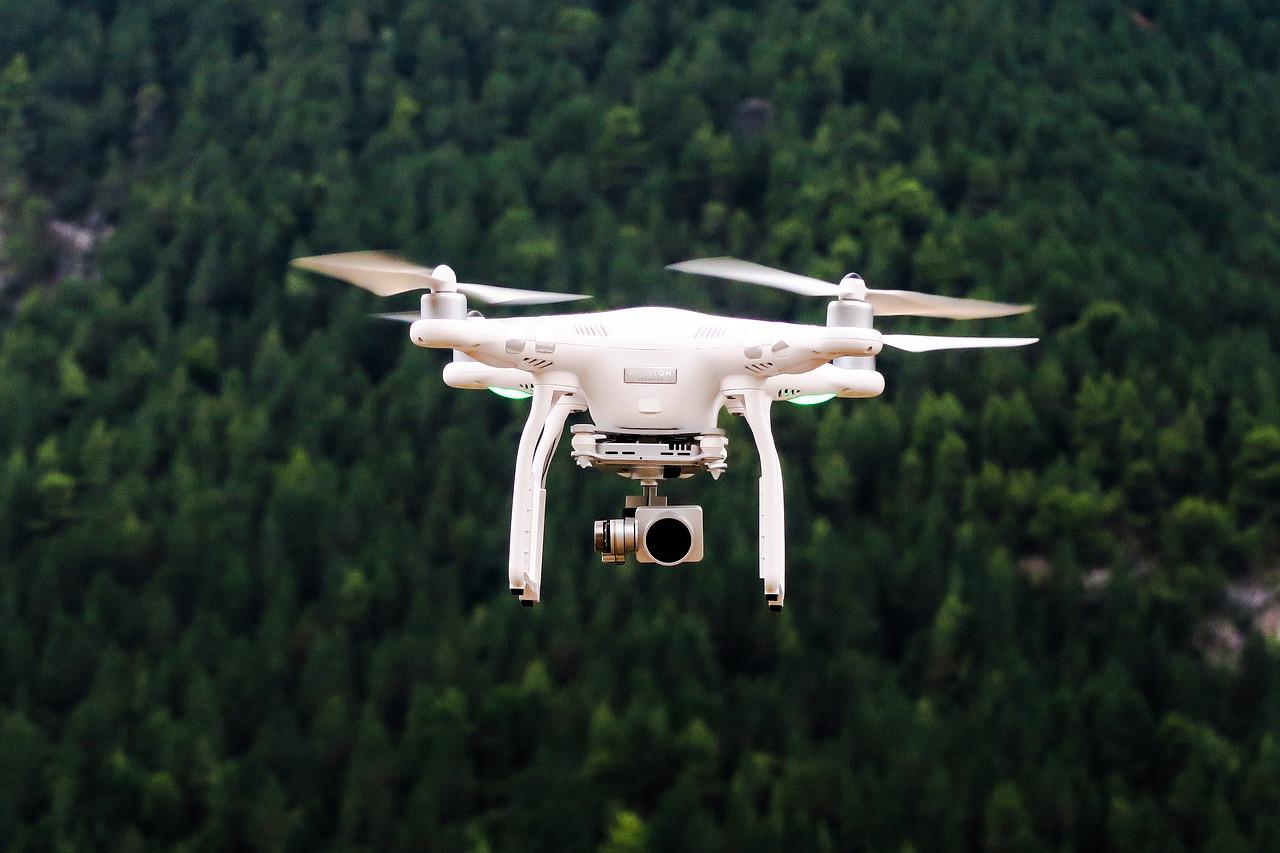 A white quadcopter drone hovering over a dark green forest
