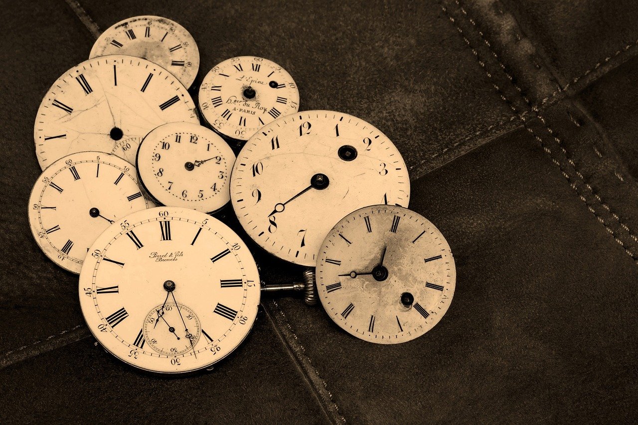 Sepia tone photo of a pile of antique clock dials set atop a canvas (or denim) background with the dials set to different times.