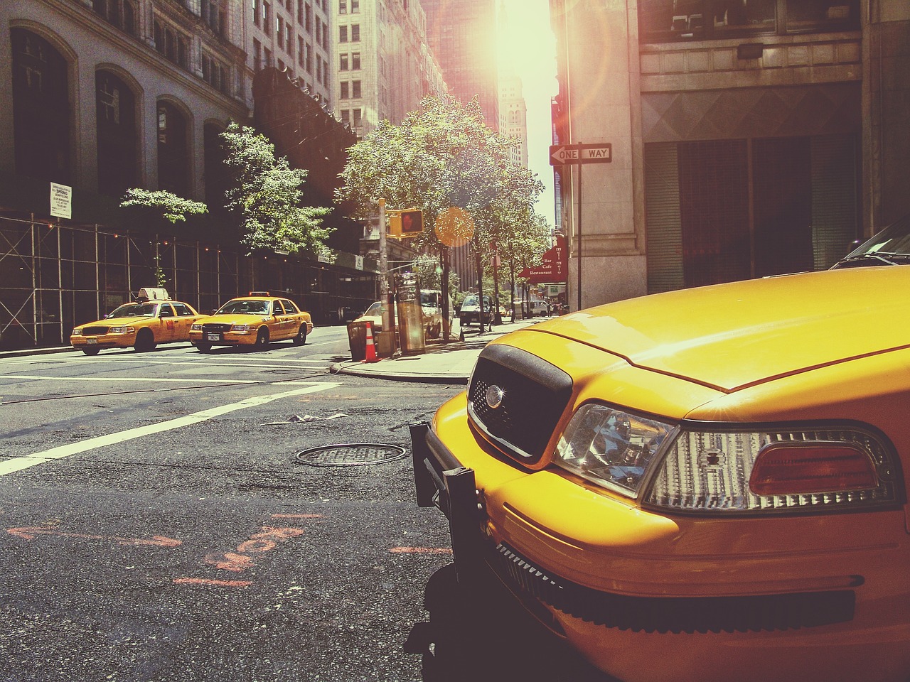 A yellow taxicab sitting on an urban city street, other taxis visible around a corner, the sun peeks brightly between skyscrapers