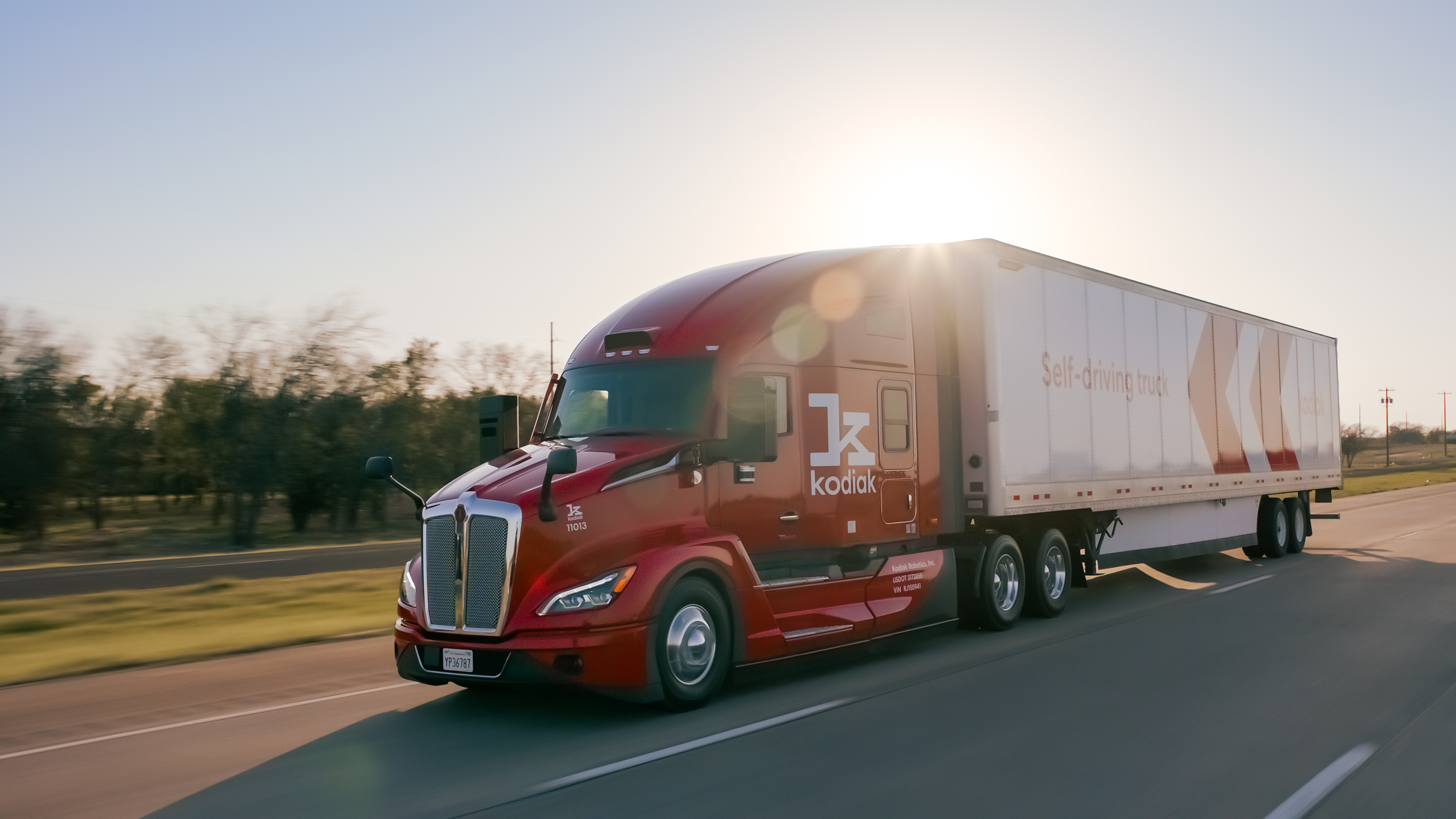 A Kodiak Robotics freight truck driving on the highway, red cab with a white cargo section, with the words in red "self-driving truck."