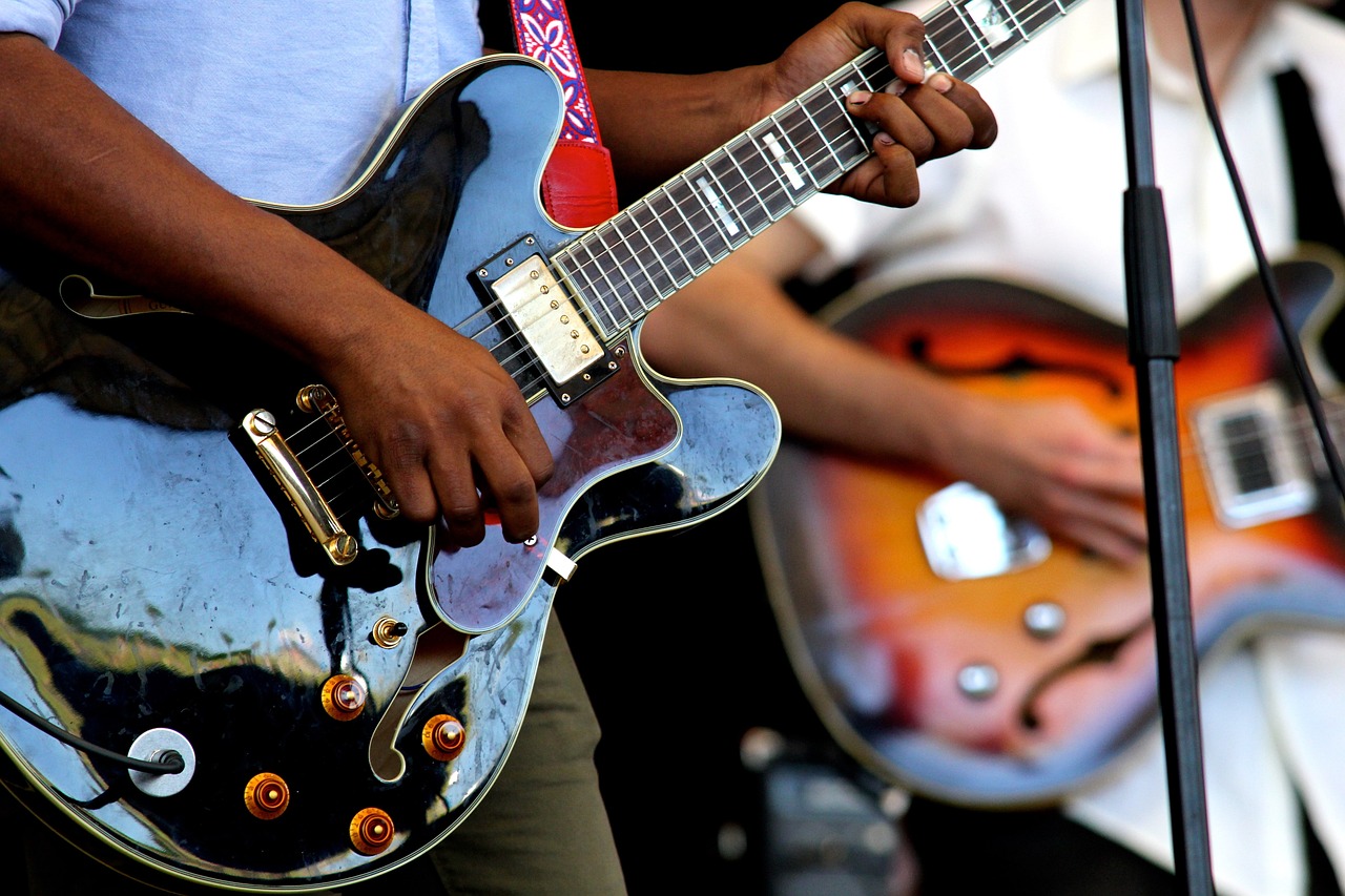 Two people holding electric guitars in front of a microphone on stage, one dark colored and one with a brown center.