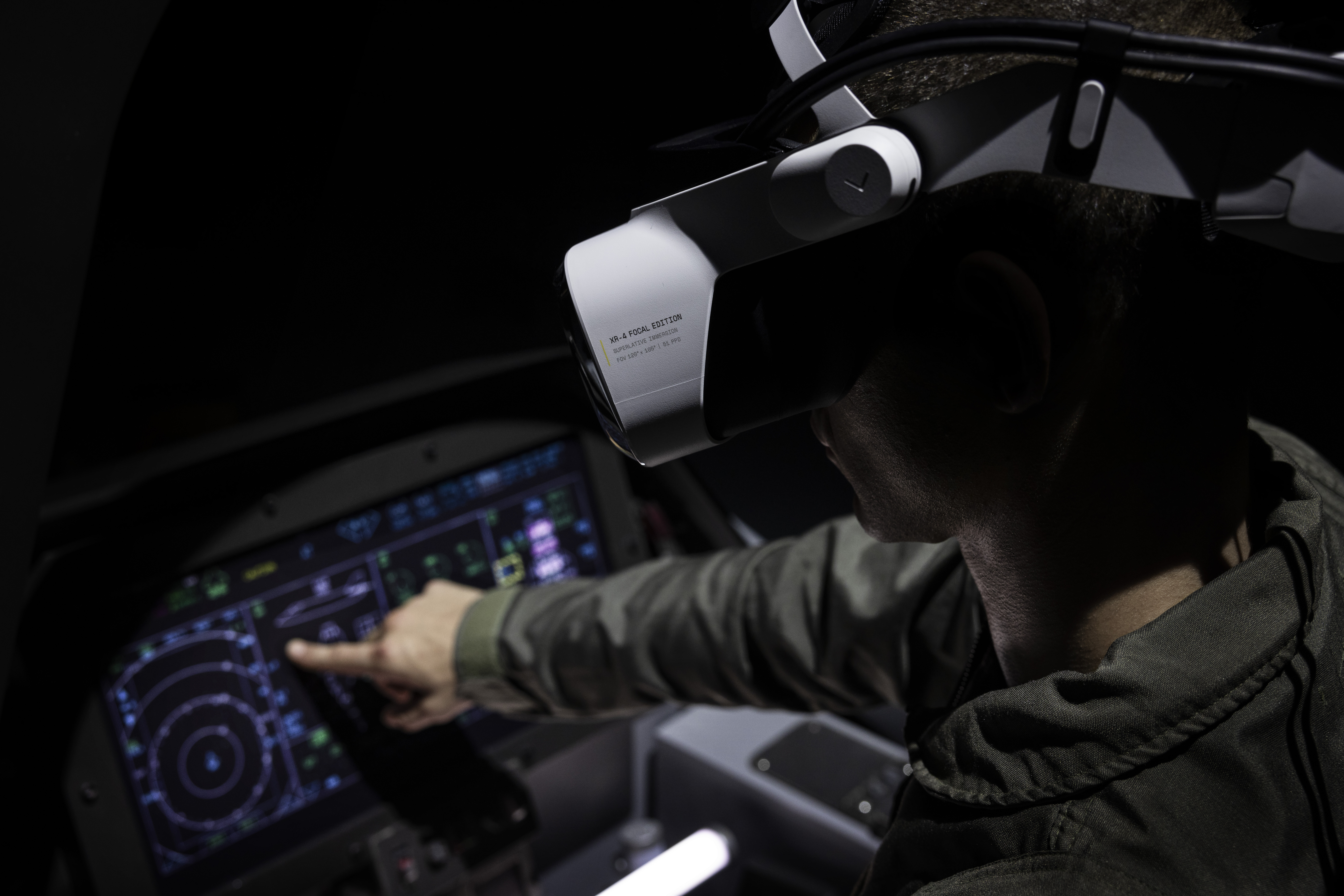 A pilot in training inside of a cockpit wearing a Varjo XR-4 Focal Edition headset, which is white and bulky on his head, the pilot has a hand reaching out towards some controls in the cockpit, which glow under an outstretched fingertip.